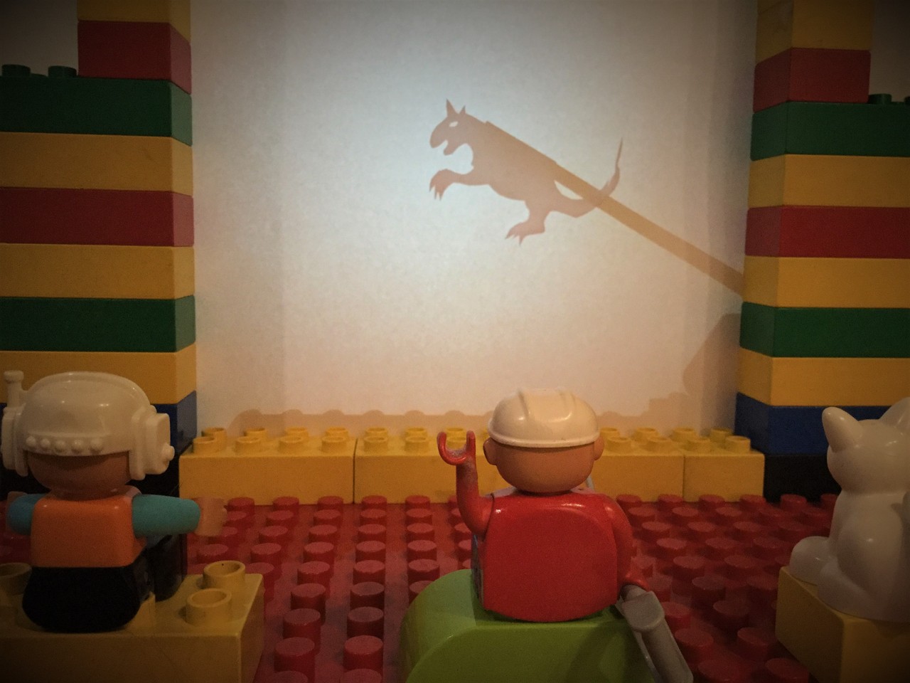 Das Foto zeigt ein Schattentheaterspiel. Ein Drache ist auf der Leinwand zu sehen. Kleine Spielfiguren beobachten das Schauspiel. Die Figur in der Mitte hebt begeistert die Hand.