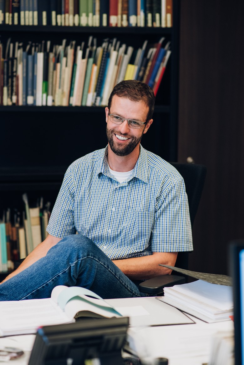 Das Foto zeigt Martin Zangl. Er sitzt in der Bibliothek vom LWL-Museum für Kunst und Kultur. Er hat sein linkes Bein über das rechte geschlagen. Er lächelt.