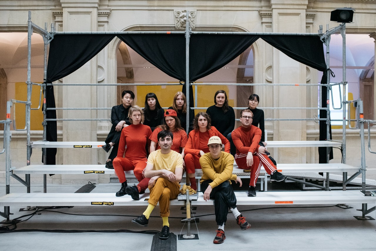 Das Foto zeigt insgesamt elf Personen. Sie alle schauen in die Kamera. Sie sitzen in drei Reihen auf einem Podest mit Stufen. Das Podest ist im Lichthof vom LWL-Museum für Kunst und Kultur. Auf der untersten und ersten Stufe sitzen zwei junge Männer. Beide sind in Gelbtönen gekleidet. Auf der zweiten und mittleren Stufe sitzen vier Personen. Drei davon sind junge Frauen. Rechts außen sitzt ein junger Mann. Sie tragen orange oder rote Kleidung. Auf der obersten und höchsten Stufe sitzen fünf Personen. Ganz links sitzt ein junger Mann. Links von ihm sitzen vier junge Frauen. Sie alle tragen schwarz.
