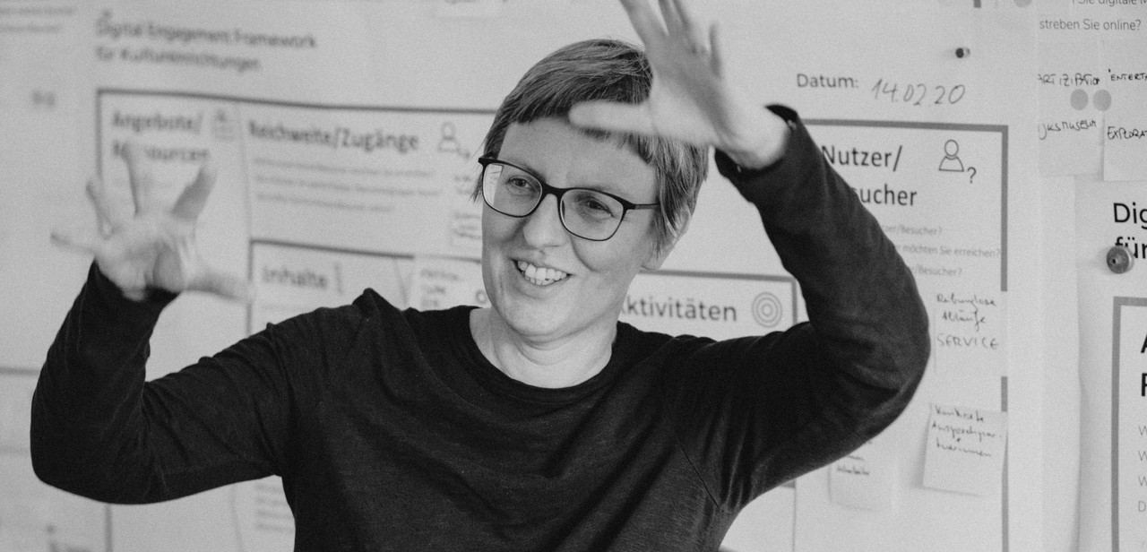 Das Foto zeigt eine Frau. Es ist Tanja Pirsig-Marshall. Sie steht vor einer Pinnwand. An die Pinnwand sind Besucherstatistiken gepinnt. Sie gestikuliert ausladend.