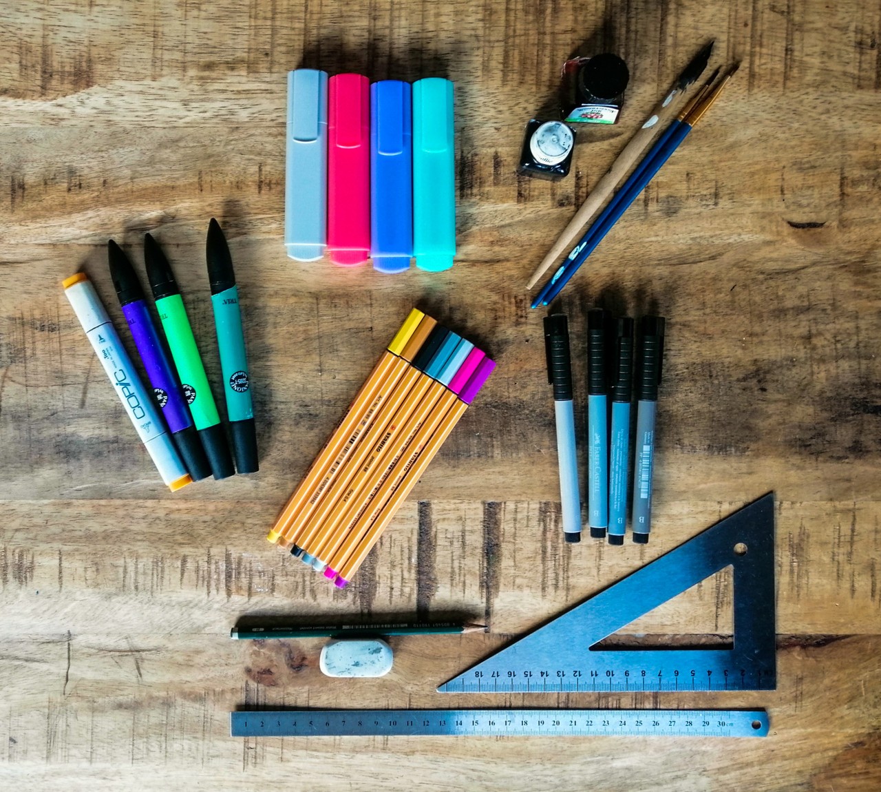 Das Foto zeigt verschiedene Schreibutensilien. Sie liegen auf einem Tisch. Man sieht Textmarker, Filzstifte, Fineliner, Füller, Pinsel und Tinte und einen Bleistift. Außerdem liegen dort ein Radiergummi, ein Lineal und ein Geodreieck.