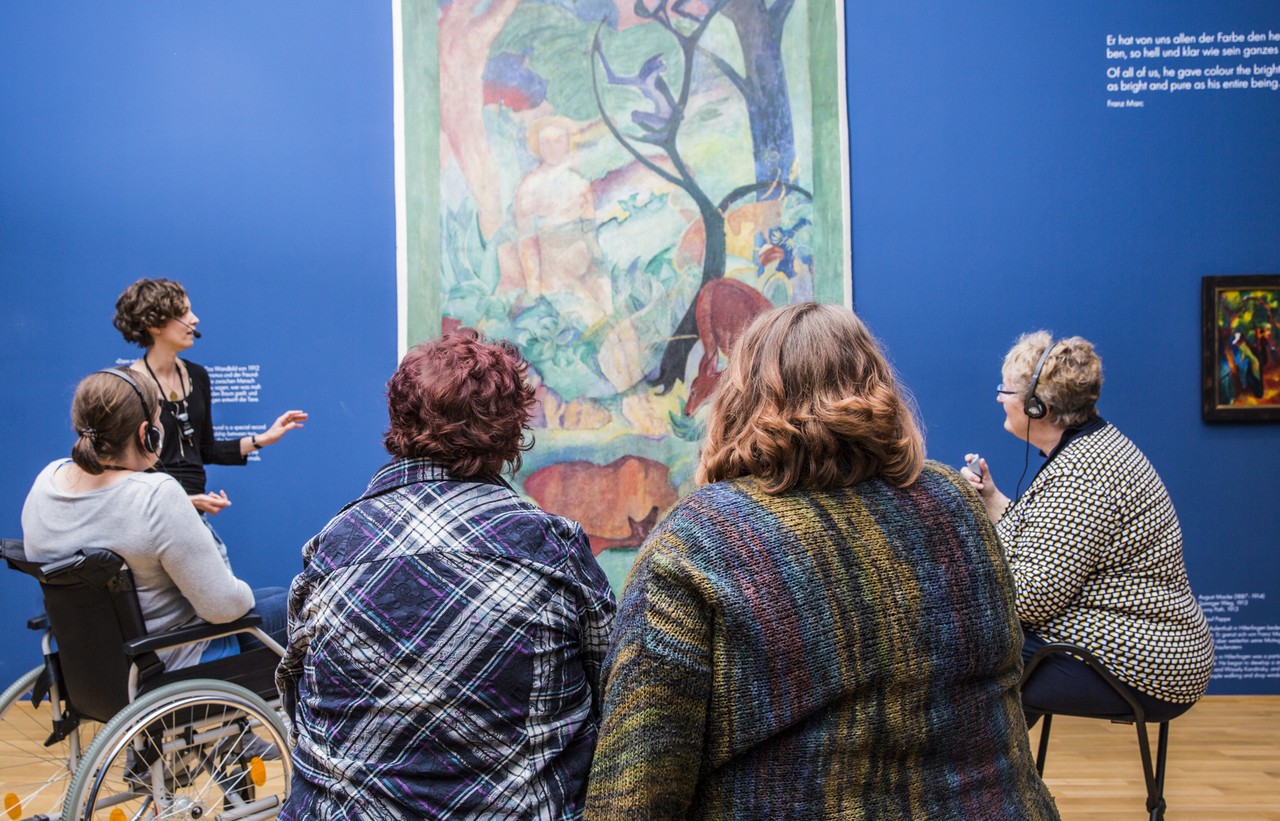 Das Foto zeigt eine Gruppe von fünf Besucher:innen bei einer Führung im LWL-Museum für Kunst und Kultur. Eine junge Frau sitzt im Rollstuhl. Eine Kunstvermittlerin erklärt ein Gemälde, das an der Wand im Hintergrund zu sehen ist.