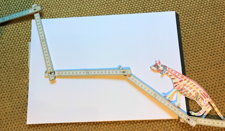 Das Foto zeigt einen Zollstock, der auf einem weißen Stück Papier liegt. Am unteren Rechten Rand von dem Bild liegt der Ausschnitt von einer gemalten Ginsterkatze.