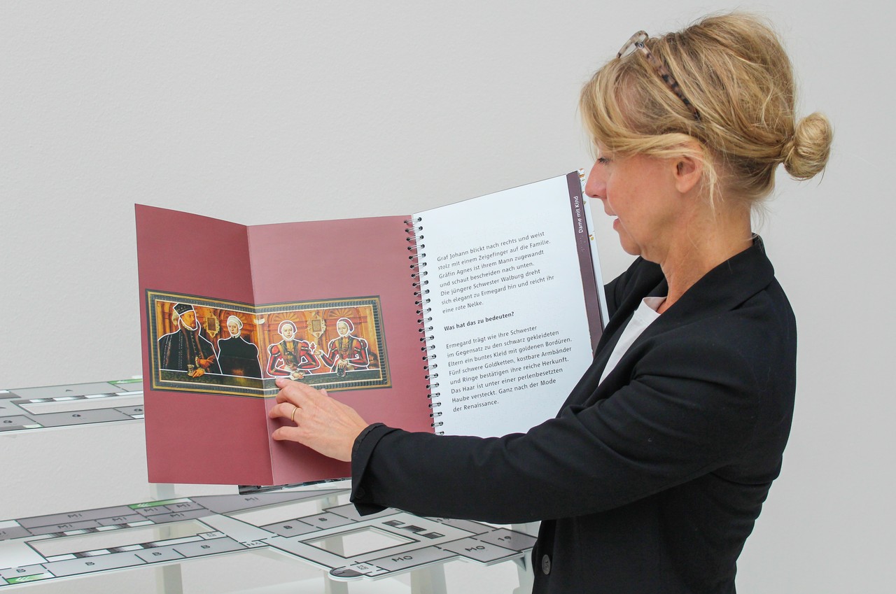 Das Foto zeigt Britta Lauro. Sie hält das Lesetastbuch vom LWL-Museum für Kunst und Kultur in den Händen. Sie zeigt darin auf das Gemälde "Familienbild des Grafen Rietberg".
