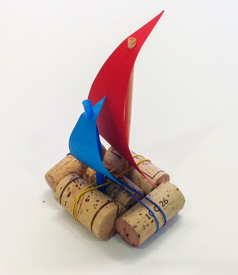 Das Foto zeigt ein kleines Boot aus Korken. Es ist mit Bindfaden zusammengebunden. Als Maste hat das Boot zwei Strohhalme. An den Strohhalmen sind jeweils Segel aus farbigem Papier befestigt.
