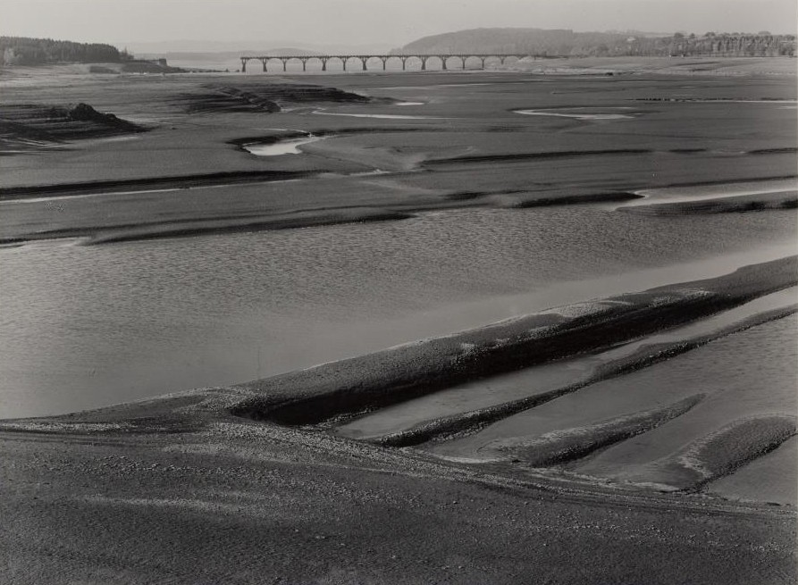 Das Foto zeigt eine graue Landschaft. Es handelt sich um eine Wattenlandschaft. Im Hintergrund kann man die Überreste einer Brücke erkennen.