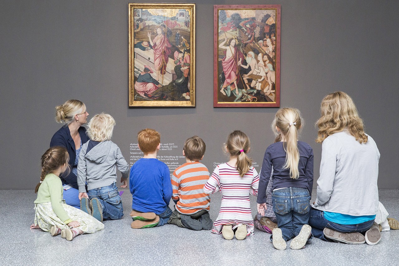 Auf dem Foto sieht man Anne Avenarius. Neben ihr ist eine Gruppe von acht Kindern und einer Frau. Alle sitzen auf dem Boden. Sie alle haben dem Betrachter den Rücken zugewandt. Sie sind in den Ausstellungsräumen vom LWL-Museum für Kunst und Kultur. Sie schauen sich zwei Gemälde an der Wand an.