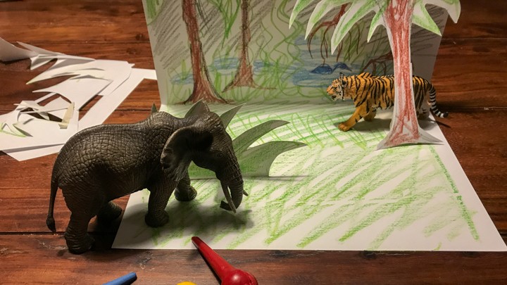 Das Foto zeigt einen Urwald als Papierkulisse. Sie steht auf einen dunklen Holztisch. Sie wurde mit Wachsmalstiften gemalt. Vor einem grünen Busch befindet sich ein Spielzeugelefant.  Hinter einer Palme steht ein Spielzeugtiger. Im Vordergrund liegen einige Wachsmalstifte.