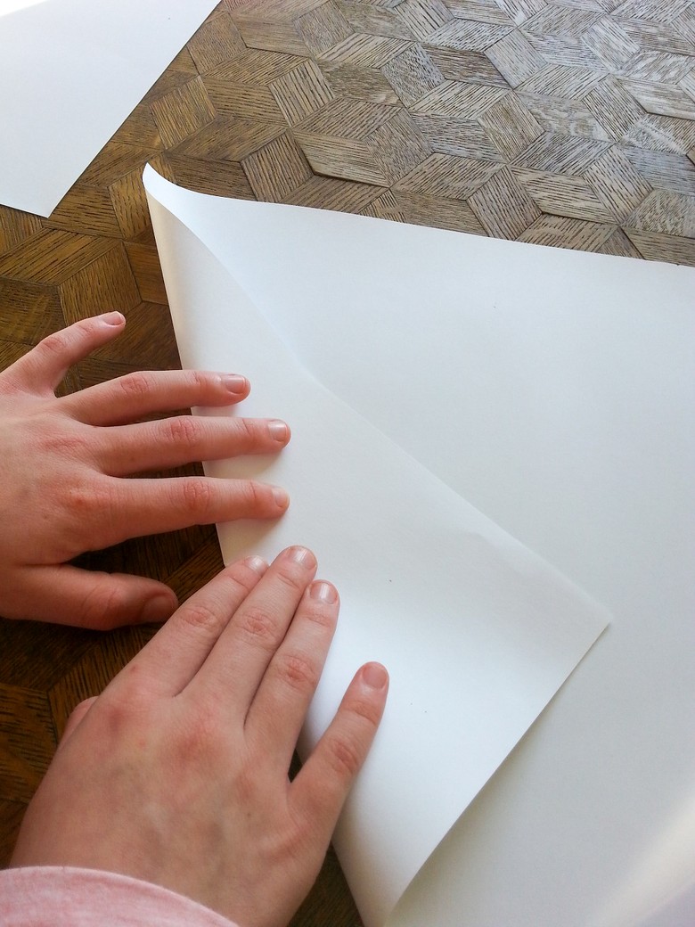 Das Foto zeigt ein Paar Hände von einer Person. Die Person faltet den unteren linken Rand von einem weißen Blatt Papier nach innen.