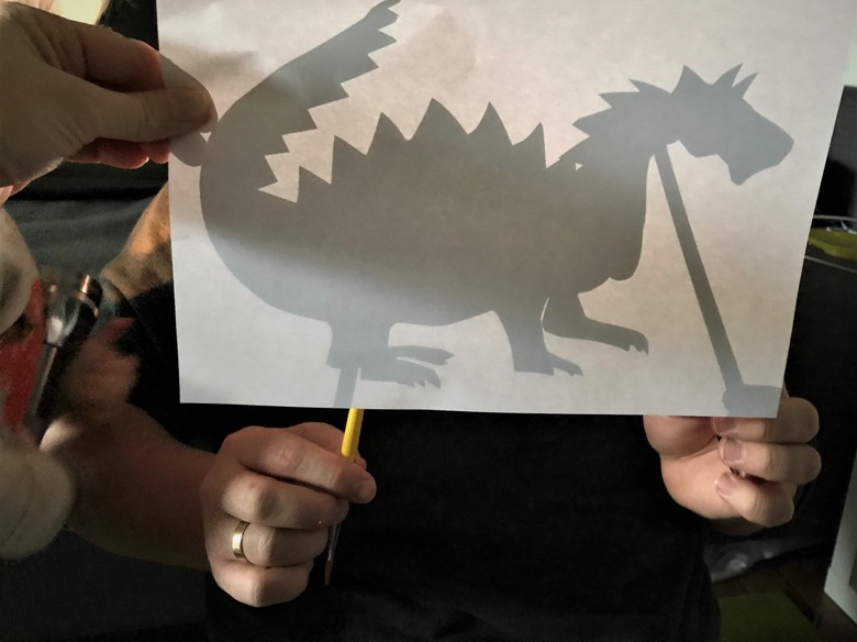 Das Foto zeigt die Silhouette eines Drachens hinter einem Blatt Papier. Es wird von der anderen Seite mit einer Lampe beleuchtet.