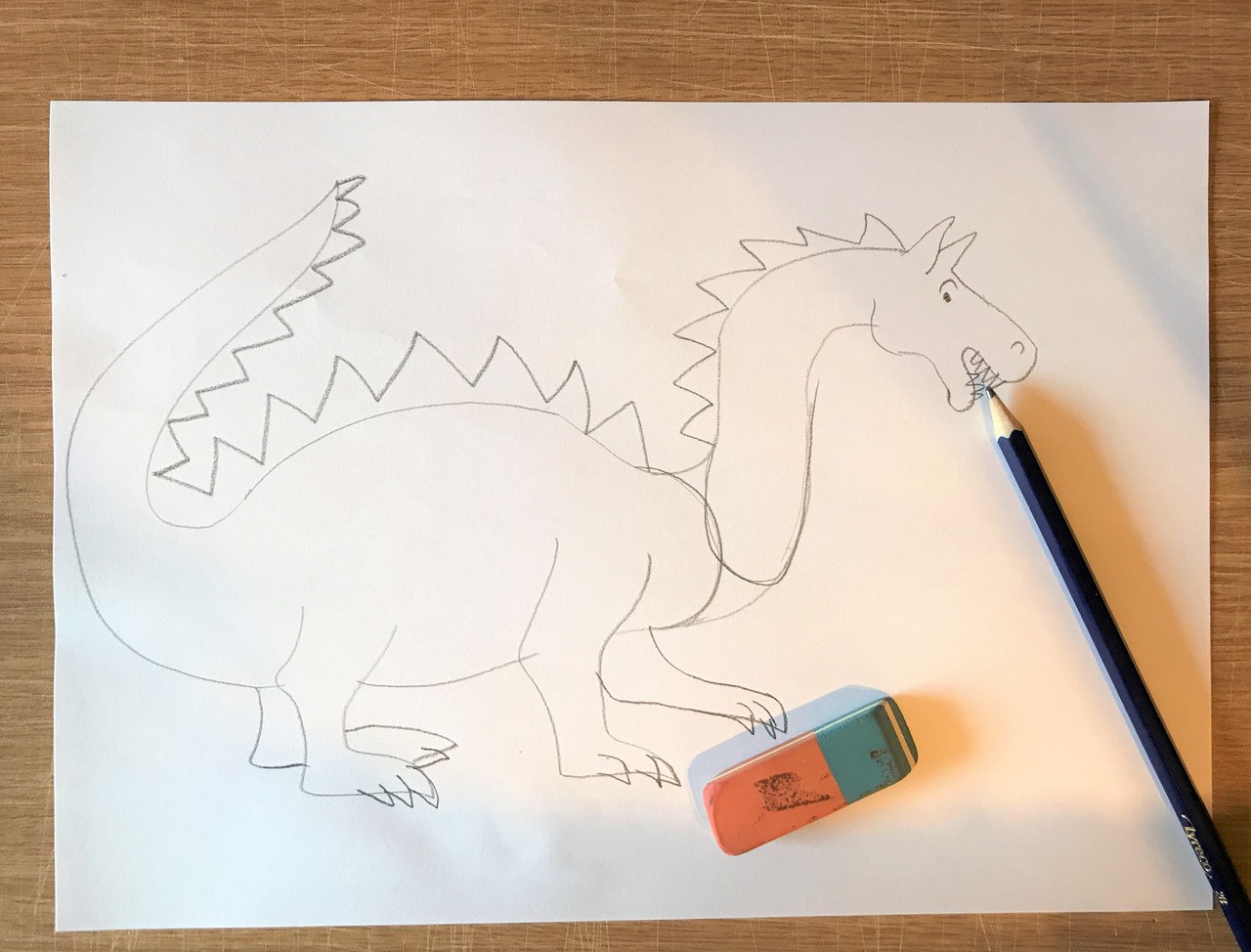 Das Foto zeigt eine Bleistiftzeichnung von einem Drachen. Er ist auf einem weißen Blatt Papier gezeichnet. Er hat einen zackigen Schuppenkamm, der sich von seinem Kopf bis zu seinem Schwanz zieht. Auf dem Blatt Papier liegen ein Bleistift und ein Radiergummi.