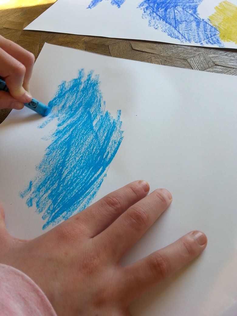 Das Foto zeigt ein Paar Hände von einer Person. Mit der linken Hand malt die Person mit einem Wachsmalstift auf einem weißen Blatt Papier. Der Wachsmalstift ist blau. Einige Linien treten deutlicher hervor. Dort wurde das Papier vorher geknickt.