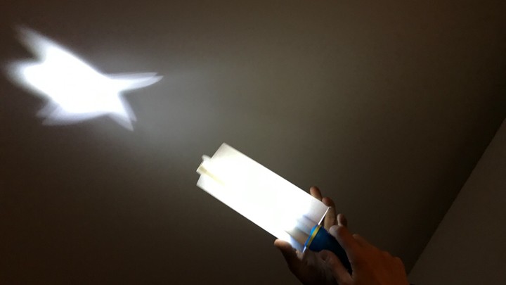 Das Foto zeigt eine Taschenlampe. Sie steckt in einer zackigen Papierröhre. Sie ist angeschaltet. Der Lichtstrahl von der Lampe ist sternförmig.