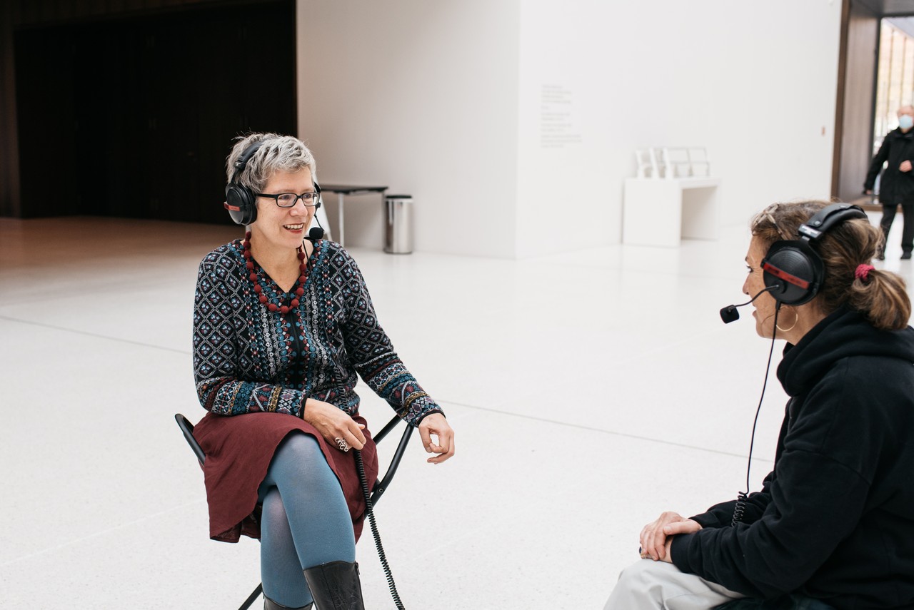 Zwei Frauen sitzen auf Hockern im weißen Foyer des LWL-Museums für Kunst und Kultur. Sie haben beide Kopfhörer auf und unterhalten sich. Im Hintergrund sieht man das Banner der Ausstellung "Passion Leidenschaft".