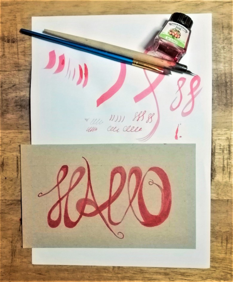 Das Foto zeigt ein Stück Papier, auf dem eine Postkarte liegt. Auf der Postkarte steht in verschnörkelter Schrift „Hallo“ in roter Tinte geschrieben. Darüber kann man auf dem Papier Schriftproben der Buchstaben sehen. Über der Schriftprobe liegen ein Pinsel, ein Füller und ein Fläschchen rote Tinte.